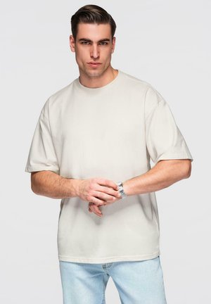 Junger Mann mit zurückgegeltem Haar, der ein schlichtes, beiges, oversized T-Shirt und hellblaue Jeans trägt und eine silberne Armbanduhr justiert.