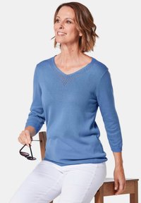 Hellblauer Strickpulli mit V-Ausschnitt und Lochmuster am Kragen. Lange Ärmel und eine taillierte Passform, kombiniert mit weißen Hosen.