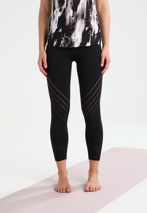 Personne debout pieds nus sur un tapis de yoga clair, portant des leggings noirs avec des découpes à motifs et un haut imprimé abstrait noir et blanc.