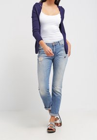 Cardigan azul marino sobre una camiseta sin mangas blanca, combinado con unos jeans desgastados de color azul claro y sandalias grises, con puños enrollados y un corte relajado.