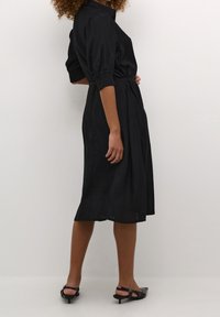 Robe noire à col montant, manches courtes bouffantes et taille ceinturée. Texture lisse, jupe jusqu'aux genoux, et fentes latérales. Chaussures à talons noires.