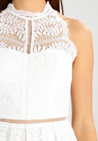 Vit spetsklänning med hög hals, prydd med detaljerade blommönster och ett genomskinligt spetstyg. Inkluderar en åtsittande liv och strukturerad tyg.