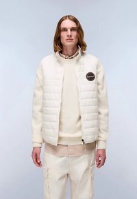 Chaqueta acolchada de color claro con cuello alto y un parche circular con logo, usada sobre un suéter crema y pantalones beige. Textura de tela lisa.