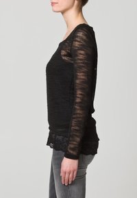 Schwarzer Strickpullover mit langen Ärmeln, mit einem strukturierten Muster und einem Spitzenabschluss. Kombiniert mit grauen Jeans, seitlich betrachtet.