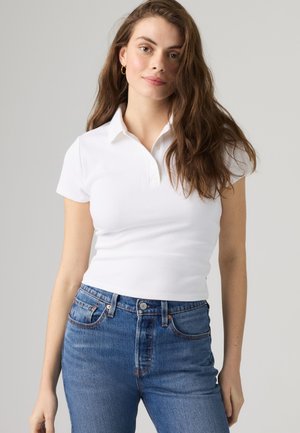 Νέα γυναίκα με μακριά καστανά μαλλιά, φορώντας ένα εφαρμοστό λευκό πόλο πουκάμισο και μπλε τζιν, στέκεται απέναντι σε ένα απλό ανοιχτό φόντο.