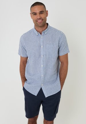 Homme souriant debout, les mains dans les poches, portant une chemise à manches courtes rayée bleu et blanc avec des boutons et un short cargo bleu marine.