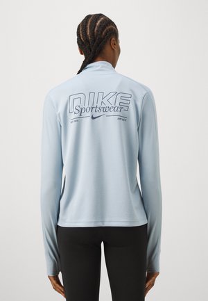 Osoba z zaplecionymi włosami, nosząca jasnoniebieską bluzę Nike Sportswear z długim rękawem i czarne spodnie, zwrócona tyłem do kamery.