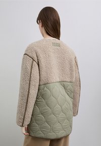 Chaqueta beige esponjosa con sección inferior acolchada de color verde oliva, con detalles acanalados y una etiqueta sutil en la parte trasera.