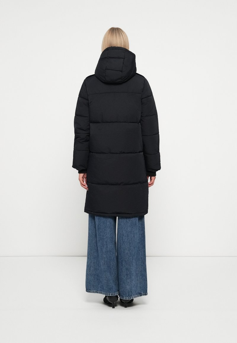 Personne blonde portant un long manteau noir matelassé avec capuche, un jean bleu à jambes larges et des chaussures noires à talons, debout, de dos.