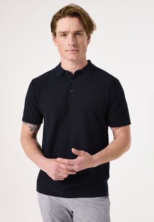 Polo bleu marine en tissu texturé, avec un col à trois boutons et des manches courtes. Il dispose d'une petite poche sur la poitrine et d'un col côtelé subtil.