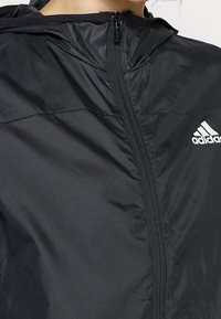 Primo piano di una persona che indossa una giacca nera Adidas con zip e cappuccio, mostrando la parte superiore del petto e il logo sul lato destro.