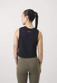 Svart kortärmad tanktop med nätdetaljer på ryggen, prydd med en gul logotyp. Tillsammans med olivgröna högmidjade leggings.