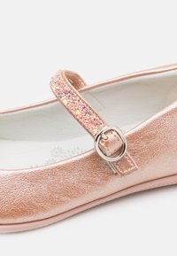 Różowozłote lakierowane buty na płaskiej podeszwie, z błyszczącym paskiem i srebrną klamrą. Wnętrze lekko wzorzyste.