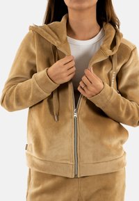 Felpa zip-up in corduroy beige con cappuccio, polsini a coste e tasche. Presenta un logo sul davanti e una texture liscia.