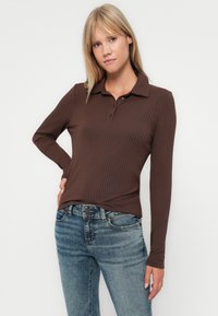 Frau trägt ein braunes, geripptes Langarm-Polohemd und blaue Jeans, steht mit einer Hand in der Hüfte vor einem einfarbigen Hintergrund.