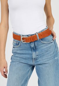 Orange kordflorsbälte med texturerad yta, med en silvermetallspänne, bärs över ljusblå högmidjade jeans och en vit linne.