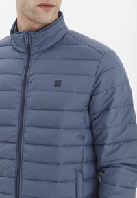 Zila pūka jaka, izgatavota no izolējoša auduma, ar augstu apkakli, rāvējslēdzēju, horizontālu šuvju un mazu logotipu uz krūšu daļas.