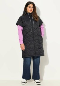 Prošívaný černý bezrukávový kabát s kapucí, se zapínáním na plný přední zip a šňůrkou. Nosený přes růžové tričko s dlouhým rukávem a džíny.