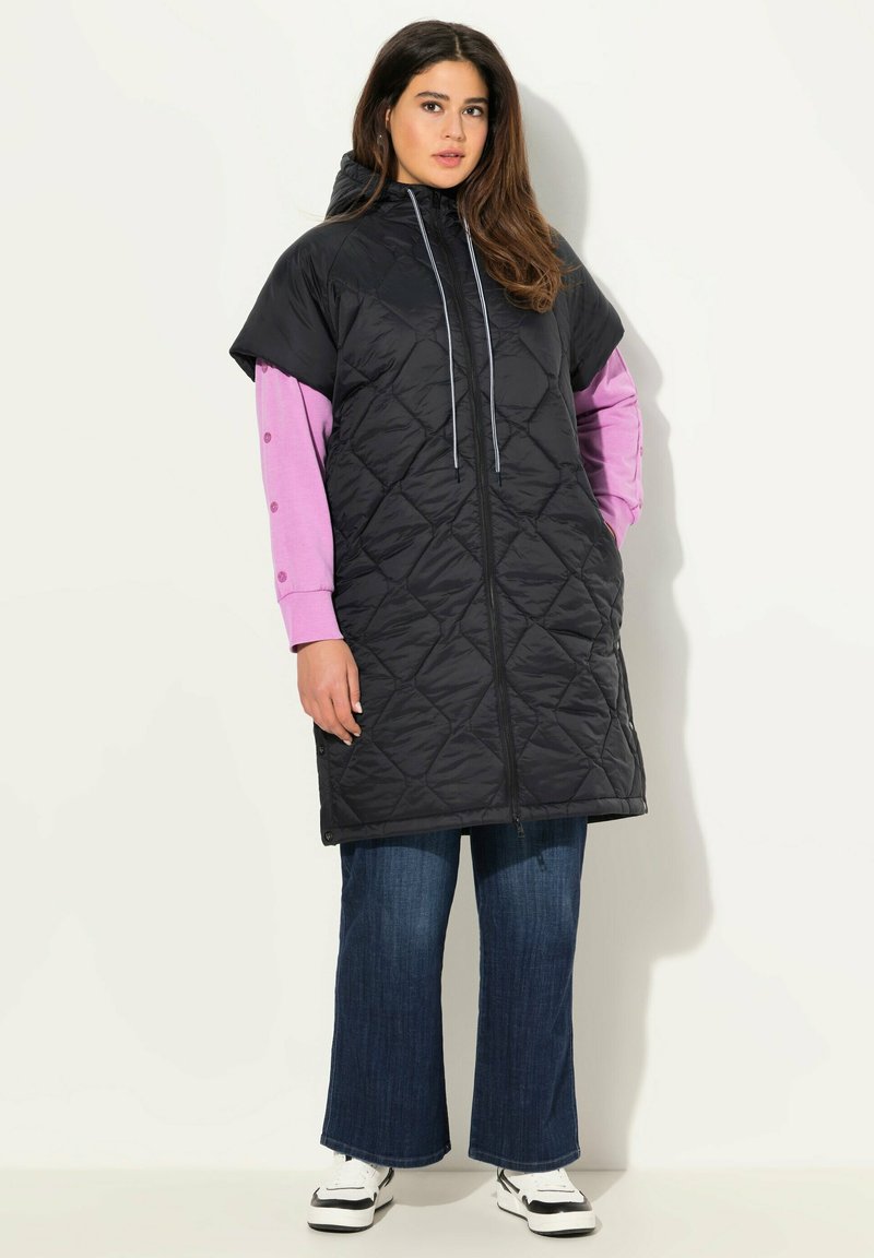 Prošívaný černý bezrukávový kabát s kapucí, se zapínáním na plný přední zip a šňůrkou. Nosený přes růžové tričko s dlouhým rukávem a džíny.