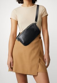 Czarna skórzana torba na ramię o eleganckim, prostokątnym kształcie i srebrnym suwakem. Nosi ją osoba w beżowej bluzce i brąskowej spódnicy z surowym dołem.