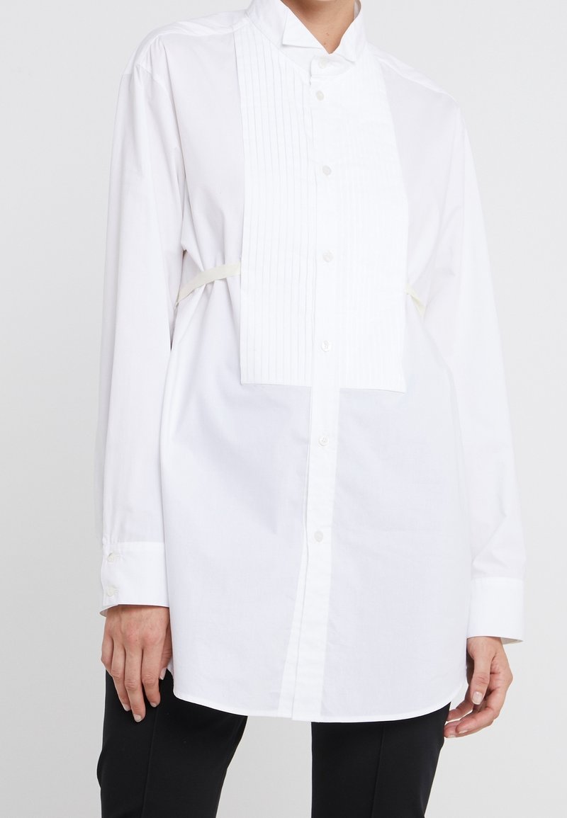 Vit skjorta med mandarin-krage och plisserad frampanel. Har långa ärmar och en avslappnad passform, tillverkad av ett mjukt tyg.
