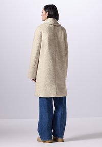 Cappotto di lana beige testurizzato con colletto alto e vestibilità ampia, abbinato a jeans blu a gamba larga e calzature chiare.