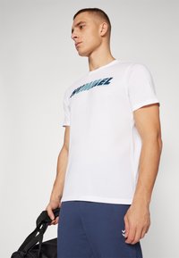 Giovane uomo con maglietta bianca Hummel e pantaloni blu che tiene una borsa da palestra nera su sfondo uniforme.