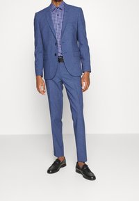 Completo blu strutturato con giacca aderente e pantaloni abbinati, abbinato a una camicia a scacchi blu e scarpe di pelle nera.
