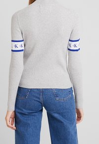 Maglione grigio a coste con maniche lunghe, caratterizzato da una banda blu e bianca su ciascuna manica con branding. Indossato con jeans in denim blu a vita alta.