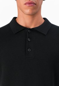 Close-up de uma pessoa vestindo uma camisola polo preta de malha com colarinho e três botões pretos na frente.