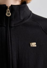 Crna zip-up jakna s visokim ovratnikom, s zlatnim metalnim logom na prsima i glatkom teksturom. Ručka za zatvarač ima ugravirane detalje.