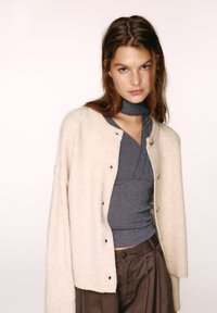 Béžový vlnený kardigán s klesnutými ramenami a čiernymi gombíkmi, vrstvený cez sivé priliehavé tričko. Hnedé voľné nohavice. Hladká textúra, ležérny štýl.