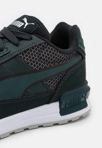 Sneaker verde scuro con pannelli in rete, sovrapposizioni testurizzate e una intersuola bianca. Include un logo Puma sulla linguetta e sul tallone.