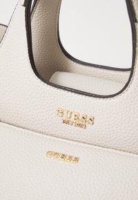 Bolso de cuero con textura crema y acentos en negro, con un logo dorado. El diseño incluye una forma estructurada y asas dobles.