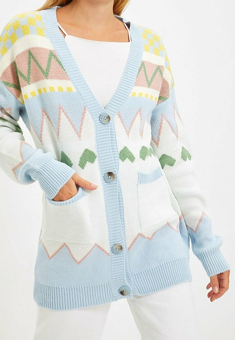 Femme portant un cardigan boutonné à motifs pastel bleu, blanc, rose et vert avec deux poches avant, sur un top blanc et un pantalon.