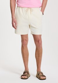 Shorts color crema con cinturilla elástica y cordón, con tejido texturizado y bolsillos laterales. Combinados con sandalias marrones.