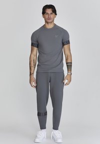 Conjunto atlético gris que presenta una camiseta ajustada de manga corta con un logo y pantalones ceñidos, acentuado por una banda de marca negra y zapatillas blancas.