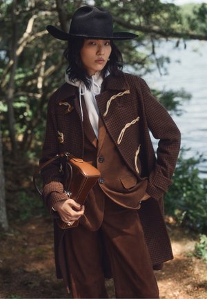 Femme porte un manteau marron à boutons à bascule par-dessus un sweat à capuche blanc et un pantalon noir, tenant un sac à main rouge et portant des lunettes de soleil foncées, debout devant un fond uni.