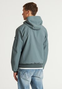 Kapuutsiga jope tuhmi teal värviga, millel on sile tekstuur ja elastne äär. Seljaosa näitab reguleeritavat kapuutsi ja küljetaskuid. Kantud heledate denim-pükstest üle.