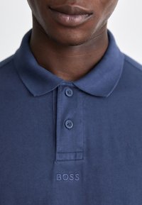 Mörkblå pikétröja i bomullstyg med krage, tre knappar och en subtil broderad logotyp "BOSS" på bröstet.