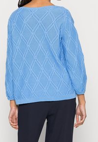 Nainen, jolla on päällään vaalean sininen neulepusero, jossa on timanttikuosi, ja mustat housut, näkyy selkäpuolelta yksinkertaista taustaa vasten.