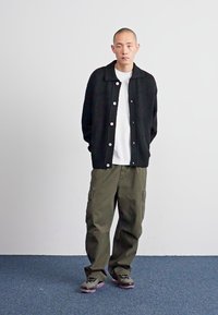 Cardigan nero a maglia con grandi bottoni bianchi, indossato sopra una t-shirt bianca. Pantaloni cargo verde oliva e sneaker grigie. Sfondo semplice.