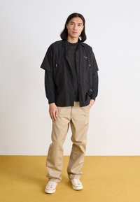 Black short-sleeve jacket with zippers, worn over a black mesh shirt, paired with beige pants and white sneakers on a yellow floor.
