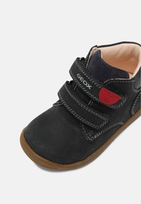 Scarpa alta in pelle nera con due cinturini in velcro, cuciture bianche a contrasto, sole in gomma e morbida fodera beige. Patcher rosso con logo sul lato.