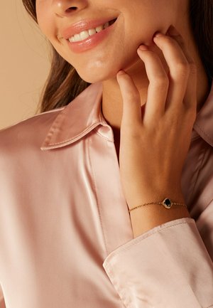Gold chain bracelet with an oval black stone centerpiece. Worn on a hand touching the face, paired with a light pink satin shirt.