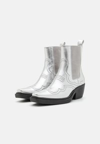Botas de tobillo de cuero plateado con punta afilada, costuras decorativas, paneles laterales elásticos y un tacón negro resistente.
