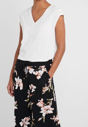 Kobieta ubrana w biały, bezrękawnik z dekoltem w serek, ozdobiony guzikami, oraz czarne kwiatowe spodnie o szerokich nogawkach z bocznymi kieszeniami.