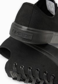 Gros plan sur des baskets en toile noire avec lacets noirs et semelle texturée portant le nom de la marque "OMBRE" sur le talon.