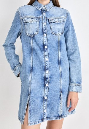 Vestido camisa de mezclilla en azul claro, con cierre frontal de botones, dos bolsillos en el pecho, mangas largas y aberturas laterales. Tela texturizada con costuras clásicas.