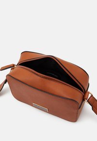Borsa a tracolla in pelle marrone con forma rettangolare, zip dorata e dettagli in piping nero. L'interno è foderato in tessuto nero, con il marchio etichettato all'interno.